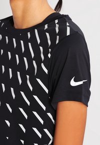 Musta urheilupaita, jossa on hopeiset vinoraitat edessä, lyhyet hihat ja pieni valkoinen Nike-logo vasemmassa hihassa.