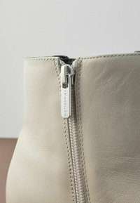 Bottine en cuir beige avec une fermeture éclair latérale et un tirant en métal argenté. Texture lisse, avec un petit talon et un design à bout arrondi.