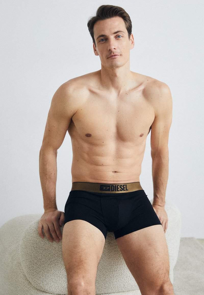 Black boxer briefs with a gold waistband featuring "DIESEL" logo. Smooth fabric, fitted design, and mid-rise cut. Model is shirtless.