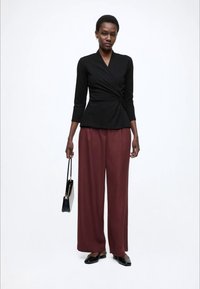 Melna apvilkta blūze ar trīs ceturtdaļu garuma piedurknēm, kopā ar vaļīgām bordo krāsas biksēm. Modeles rokās ir maza melna rokassomiņa, un viņa valkā melnas dzīvokļapavus.