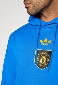 Modrá mikina s textúrovaným povrchom, so čierno-zlatým logom Manchester United a žltým emblémom Adidas trefoil nad kapucňou.