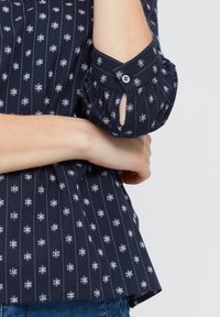 Camicia blu con bottoni, caratterizzata da strisce verticali bianche e piccoli motivi geometrici, con dettagli di polsini arricciati e una texture del tessuto morbida.