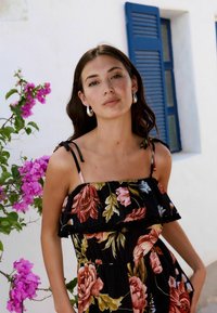Kvetinové letné šaty z čierneho materiálu s volánovými ramienkami, zdobené farebnými ružami a listami. Strieborné náušnice v tvare kruhu dopĺňajú tento outfit.