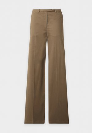 Femme debout, portant une chemise foncée à boutons, un pantalon beige large et tenant un sac à main noir, faisant face à l'avant dans un studio sobre.