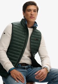 Groene pufferbodywarmer met verticale stiksels, gedragen over een crèmekleurige textuursweater. Blauwe geruite overhemdmanchetten zichtbaar, gecombineerd met denim jeans.