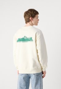 Felpa color crema con una grafica testurizzata verde sul retro che presenta montagne stilizzate e testo. Polsini e orlo a coste. Vestibilità casual.