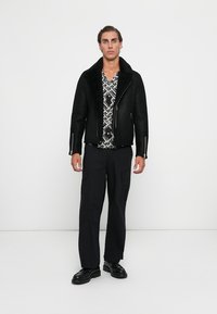 Hombre de pie con chaqueta de piel de oveja negra, pantalones cargo negros, mocasines negros y una camisa con patrón en blanco y negro, contra un fondo blanco.