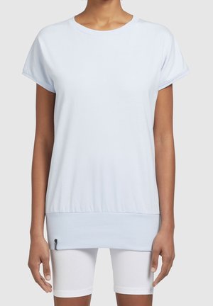 Persoon die een lichtblauw T-shirt met korte mouwen draagt, met een brede geribde zoom en witte aansluitende korte broek tegen een effen achtergrond.