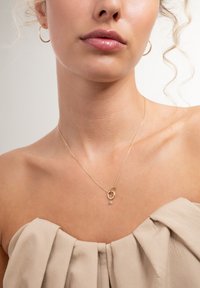Woman wearing gold hoop earrings and a delicate gold necklace with two interlocking rings and a small hanging gemstone.