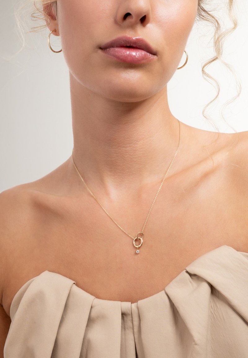 Woman wearing gold hoop earrings and a delicate gold necklace with two interlocking rings and a small hanging gemstone.