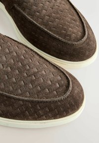 Chaussures en daim marron sans lacets avec un motif texturé en forme de diamant et des semelles en caoutchouc blanc, présentées en gros plan sur un fond clair.