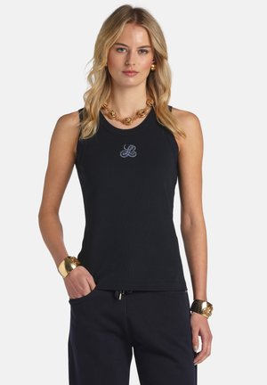 Mujer rubia con top negro sin mangas con un pequeño logo bordado, pantalones negros, collar de cadena dorada y pulseras doradas a juego.