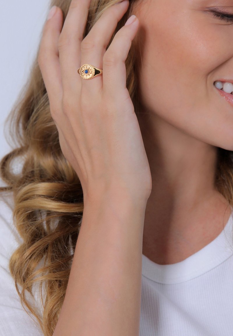 Bague en or avec une pierre précieuse bleue centrale, bande texturée, portée sur une main avec des cheveux doux et ondulés. Vêtements blancs à texture côtelée.
