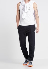 T-shirt blanc sans manches avec un logo, pantalon de survêtement marine, et baskets sportives avec semelles orange. La tenue comprend un haut à capuche posé sur les épaules.