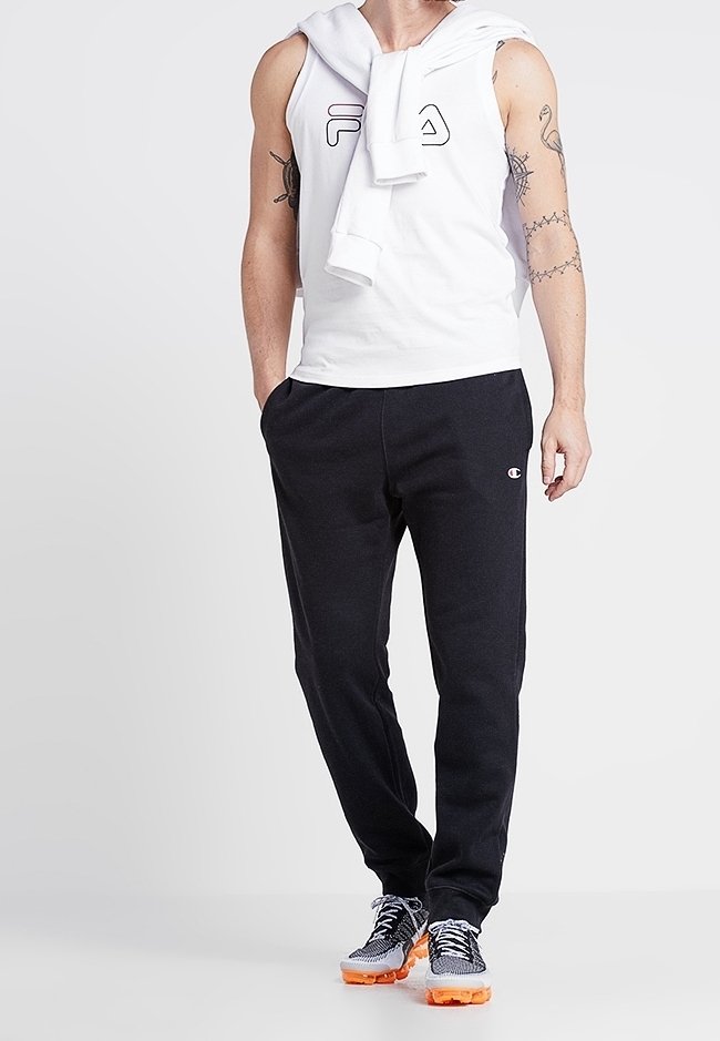 T-shirt blanc sans manches avec un logo, pantalon de survêtement marine, et baskets sportives avec semelles orange. La tenue comprend un haut à capuche posé sur les épaules.