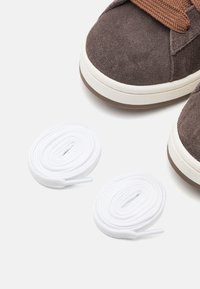 Bruna mockasneakers med vita gummisulor och bruna skosnören; två extra vita skosnören i rullar bredvid skorna.