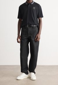 Zwarte polo met grijze accenten, voorzien van een logo. Donkerdenim broek met een relaxed fit en witte sneakers. Eenvoudig en schoon ontwerp.