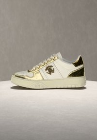 Baskets blanches basses avec des accents dorés, logo sur le côté, talon texturé et semelle en caoutchouc crème sur fond neutre.