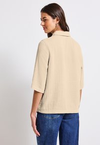 Camicetta beige a maniche corte realizzata in tessuto testurizzato, con colletto, dettaglio con apertura sulla schiena e vestibilità leggermente ampia, abbinata a jeans blu.