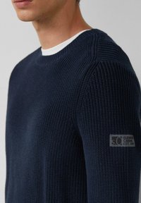 Marineblau gestrickter Pullover mit gerippter Textur, Rundhalsausschnitt und einem grauen Logoschild auf dem linken Ärmel. Verfügt über eine enganliegende Passform.