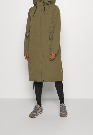 Kobieta w oliwkowo-zielonej długiej pikowanej kurtce z kapturem, czarnych legginsach i szarych tenisówkach, stojąca na jasnym, jednolitym tle.