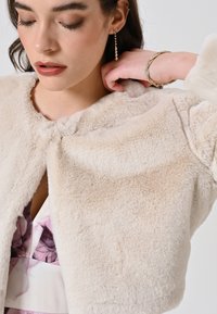 Veste courte en fausse fourrure de couleur beige clair, avec une texture douce et un col rond. Présente un fermoir à l'avant et une vue latérale d'une robe à fleurs en dessous.