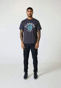 Man standing facing forward wearing a black graphic t-shirt, black cargo pants, and black sneakers against a plain background.