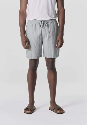 Osoba ubrana w jasnoszare szorty z troczkiem, białą koszulkę bez rękawów i brązowe klapki na płaskiej podeszwie, stojąca na jednolitym tle.