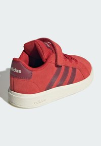 Crvena Adidas tenisica s čičak trakom, tri tamnocrvene pruge i bijeli potplat, lagano nagnuta kako bi se prikazala stražnja i bočna strana.