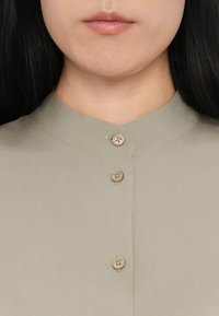 Light olive green blouse with a high neckline and four brown buttons. Features smooth fabric with minimal texture and clean stitching.