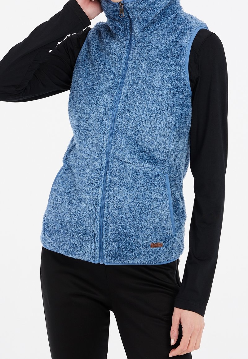 Kvinna iklädd en blå fluffig zip-up ärmlös väst över en svart långärmad topp och svarta byxor, med en hand som rör vid hennes huvud.