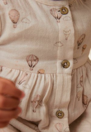 Jasno beżowa sukienka z powtarzającym się wzorem balonów na gorące powietrze w delikatnych kolorach. Posiada marszczoną talię i drewniane guziki na przodzie.