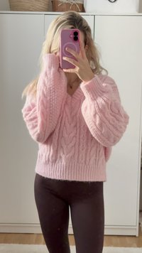 Svetr s kabelkovým pletením a výstřihem do V v jemně růžové barvě; má oversized rukávy a žebrovaný lem. Kombinován s tmavými legínami na neutrálním pozadí.