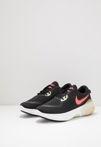 Zapatillas deportivas negras con una parte superior de malla, detalles en rosa y una suela blanca con una sección del talón translúcida. Perforaciones circulares para la transpirabilidad.