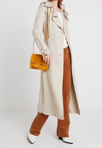 Beige trenchcoat met een gekartelde kraag, knoopsluiting aan de voorkant en zijzakken; gecombineerd met rustkleurige corduroypantalons en een gele handtas.
