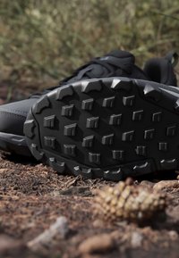 Chaussure d'extérieur noire avec une semelle épaisse et robuste présentant des motifs quadrangulaires et angulaires sur un sol forestier avec des pommes de pin et des brindilles.