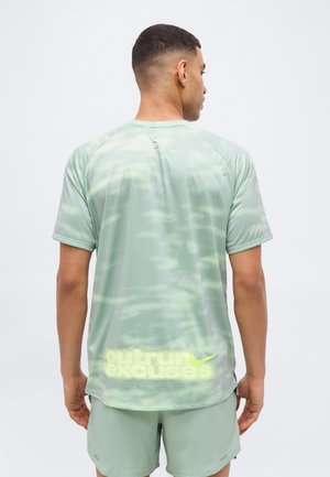 Άνδρας που φοράει αθλητική μπλούζα και σορτς σε μέντα πράσινο χρώμα, με το κείμενο "outrun excuses" και το λογότυπο της Nike κοντά στο κάτω μέρος της πλάτης, κοιτάζοντας προς τα μακριά από την κάμερα.