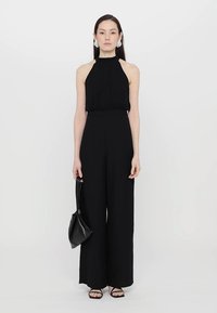 Svart halterneck jumpsuit med vide ben, glat stof og tætsiddende bodice. Sammen med en lille sort taske og sandaler med remme.