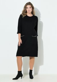 Schwarzes, gestricktes Kleid mit 3/4-Ärmeln, einem Rundhalsausschnitt und Taschenakzenten, die mit weißen Perlen verziert sind. Glatte Textur, knielange Passform.