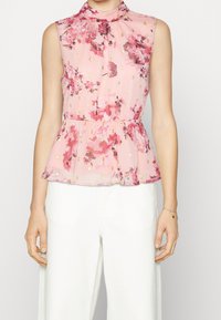 Bledě růžová květinová peplum top s vysokým výstřihem a bezramenným designem. Má zlaté akcenty a lehkou, průsvitnou látku. Nošeno s bílými kalhotami.