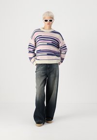 Strikkegenser med lilla og rosa striper, rund hals, kombinert med løse, mørke denimjeans og svarte sko. Modellen bærer hvite solbriller.