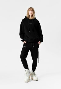 Sudadera negra con cordones ajustables y detalles en blanco; combinada con pantalones jogger negros que tienen un bolsillo con cremallera y detalles en blanco. Botas de combate blancas.