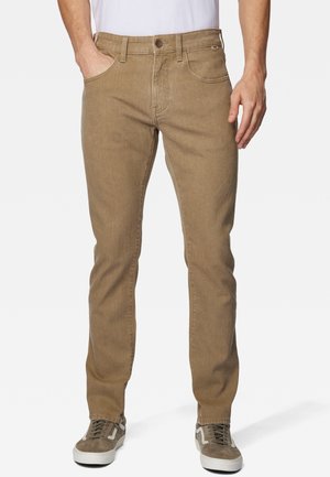 Homme portant un pantalon beige slim, un t-shirt blanc et des baskets marron aux semelles blanches, debout avec une main dans la poche.