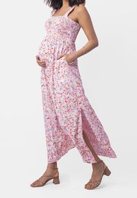 Femme enceinte portant une robe maxi fleurie rose avec une fente sur le côté et des sandales à talon bloc brunes à lanières, tenant son ventre.