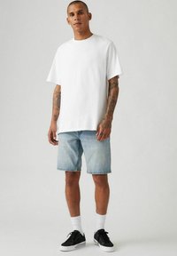 Weißes Baumwoll-T-Shirt mit kurzen Ärmeln, kombiniert mit hellblauen Denim-Shorts. Schwarze Sneakers und weiße Socken vervollständigen das Outfit.