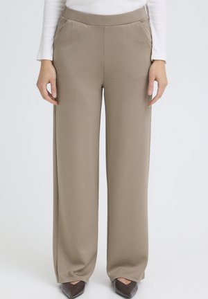 Pantaloni beige a gamba larga con cintura elastica indossati con una maglia bianca a maniche lunghe e scarpe marroni con punta, mani rilassate lungo i fianchi.