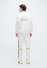 Άνδρας με την πλάτη γυρισμένη, φορώντας λευκή φόρμα με μαύρες ρίγες και πράσινο λογότυπο κροκοδείλου στο πίσω μέρος του μπουφάν.