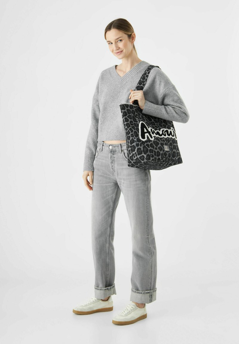 Femme portant un pull gris et un jean, tenant un sac fourre-tout à motif léopard noir avec lettrage blanc sur l'épaule, se tenant sur fond blanc.