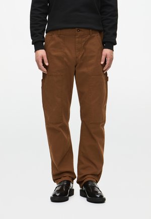 Hnedé cargo nohavice z bavlny, s bočnými vreckami, vo voľnom strihu a s kontrastným šitím. V kombinácii s čiernymi topánkami.