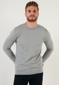 Maglione grigio chiaro con motivo a nido d'ape, collo rotondo, polsini e orlo a costine. Indossato con jeans blu scuro.